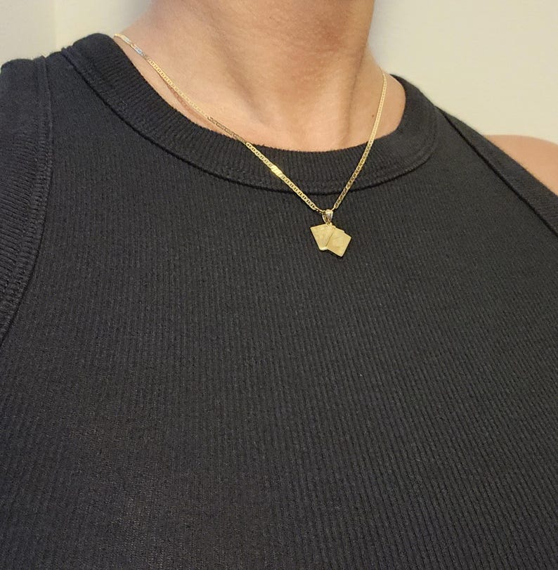 May include: A gold-toned necklace featuring a delicate chain and a small pendant shaped like two playing cards. The pendant hangs from a simple bail, and the entire piece is worn against a black ribbed sleeveless top, highlighting the metallic finish.