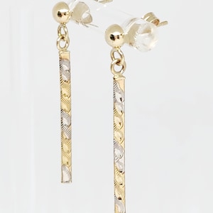 May include: A pair of gold dangle earrings with a two-tone design. Each earring features a small gold ball at the top, connected to a long, rectangular bar with alternating gold and silver-colored patterns. The earrings are displayed against a white background.