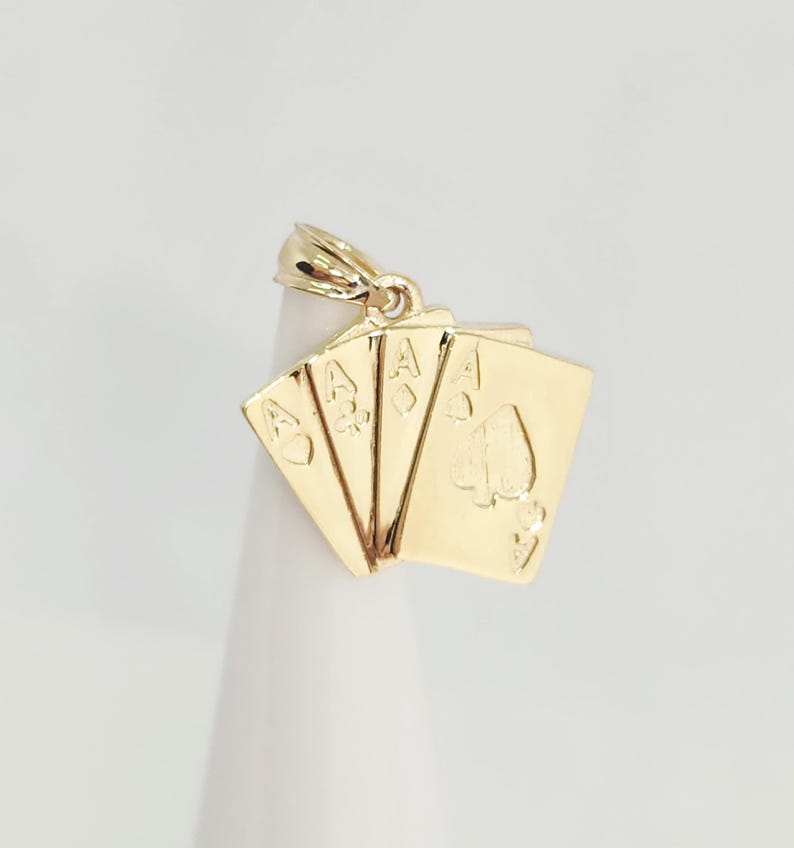 May include: A gold pendant shaped like a fan of four playing cards, each featuring an ace. The cards display the heart, club, diamond, and spade suits. The pendant includes a small gold bail at the top for attachment to a chain or necklace.
