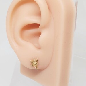 Puede incluir: Un pendiente de bot&oacute;n dorado con forma de hada se muestra en una oreja de maniqu&iacute;. El hada tiene alas extendidas y lleva un sombrero. El pendiente es peque&ntilde;o y detallado, con rasgos faciales y ropa visibles.