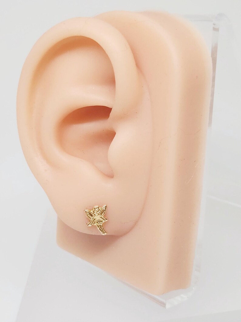 Puede incluir: Un pendiente de bot&oacute;n dorado con forma de hada se muestra en una oreja de maniqu&iacute;. El hada tiene alas extendidas y lleva un sombrero. El pendiente es peque&ntilde;o y detallado, con rasgos faciales y ropa visibles.
