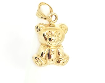 May include: A polished gold pendant shaped like a seated teddy bear. The charm features a smooth, reflective surface with defined ears, a rounded snout, and small paws. A small, circular gold bail is attached to the top for hanging on a chain.