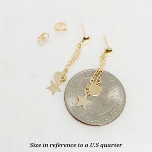May include: Gold dangle earrings with a star and heart charm. The earrings are shown next to a US quarter for size reference.
