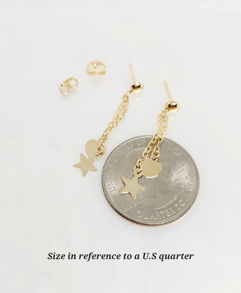 May include: Gold dangle earrings with a star and heart charm. The earrings are shown next to a US quarter for size reference.