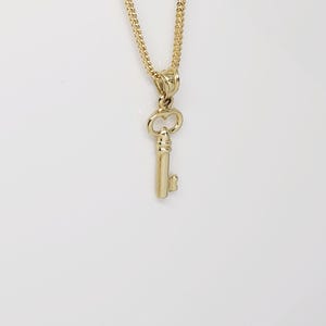 May include: A gold-colored key pendant necklace. The key charm is detailed with a classic key shape and is suspended from a delicate chain. The necklace is set against a white background.