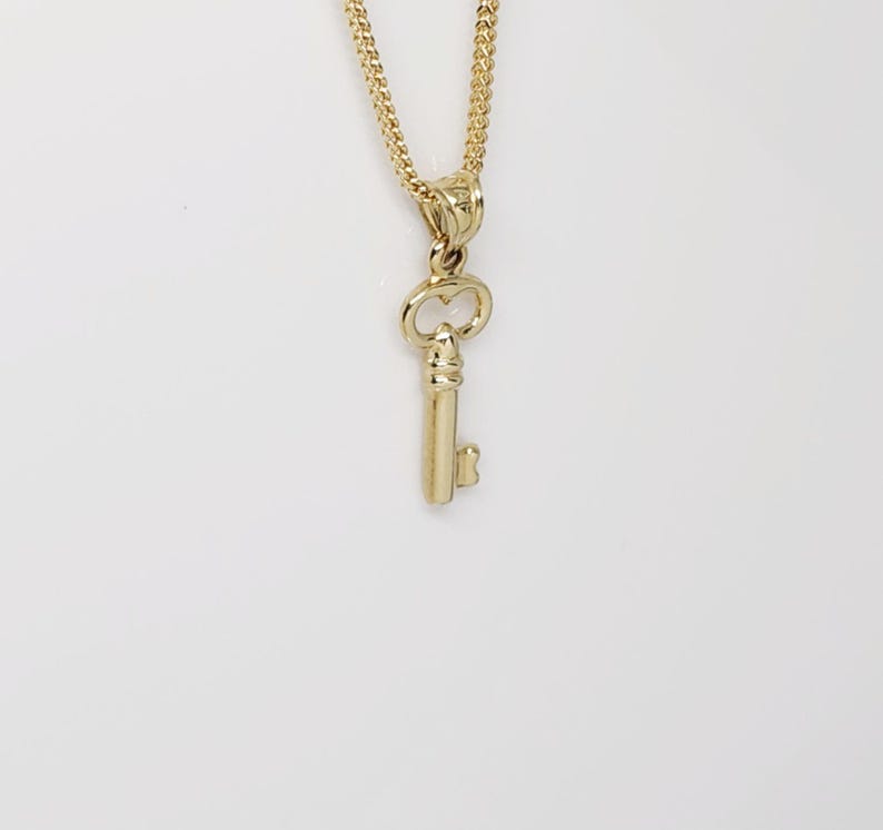 May include: A gold-colored key pendant necklace. The key charm is detailed with a classic key shape and is suspended from a delicate chain. The necklace is set against a white background.