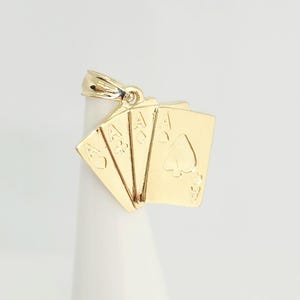 May include: A gold pendant shaped like a fan of four playing cards, specifically the ace of hearts, ace of clubs, ace of diamonds, and ace of spades. The metal piece features a polished finish and includes a small bail at the top for attachment.