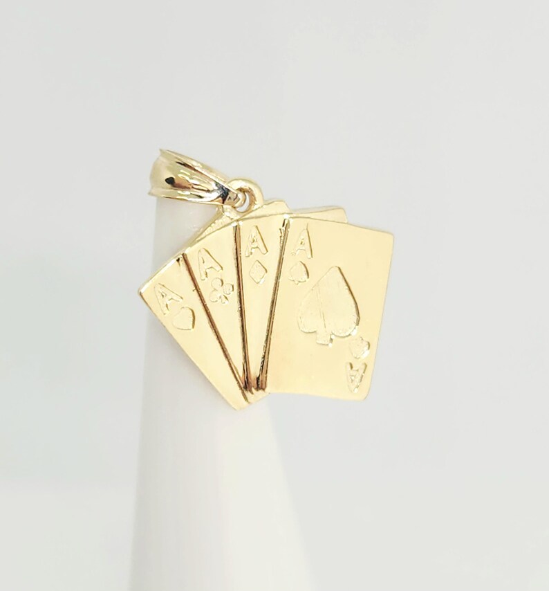 May include: A gold pendant shaped like a fan of four playing cards, specifically the ace of hearts, ace of clubs, ace of diamonds, and ace of spades. The metal piece features a polished finish and includes a small bail at the top for attachment.