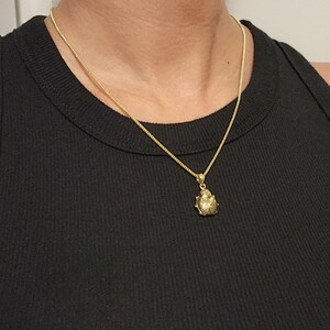 K&ouml;nnte beinhalten: Eine goldfarbene Halskette mit einer zarten Kette und einem kleinen, strukturierten Anh&auml;nger in Form eines Marienk&auml;fers. Der Anh&auml;nger hat eine abgerundete, metallische Oberfl&auml;che mit feinen Gravuren und h&auml;ngt an einer einfachen, d&uuml;nnen Kette.