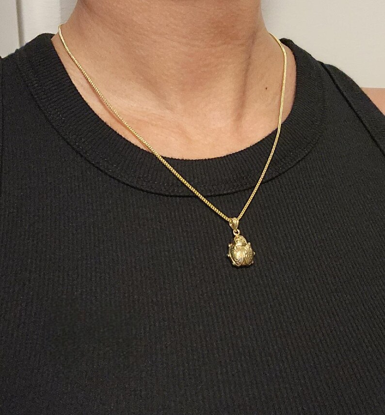 K&ouml;nnte beinhalten: Eine goldfarbene Halskette mit einer zarten Kette und einem kleinen, strukturierten Anh&auml;nger in Form eines Marienk&auml;fers. Der Anh&auml;nger hat eine abgerundete, metallische Oberfl&auml;che mit feinen Gravuren und h&auml;ngt an einer einfachen, d&uuml;nnen Kette.
