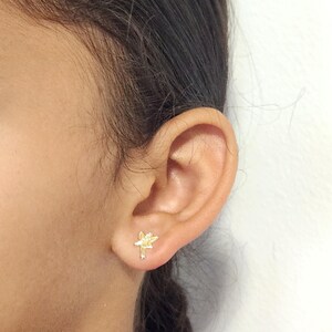 Puede incluir: Primer plano de una oreja con un peque&ntilde;o pendiente de bot&oacute;n con forma de flor dorada. El pendiente est&aacute; detallado con p&eacute;talos y un tallo. El fondo es una pared blanca lisa.