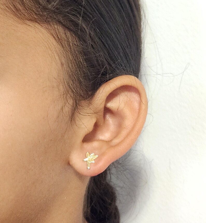 Puede incluir: Primer plano de una oreja con un peque&ntilde;o pendiente de bot&oacute;n con forma de flor dorada. El pendiente est&aacute; detallado con p&eacute;talos y un tallo. El fondo es una pared blanca lisa.