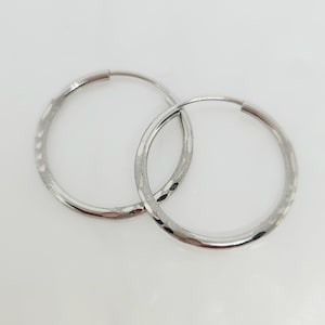May include: A pair of silver-toned hoop earrings featuring a polished finish with subtle, repeating etched indentations along the outer surface of the circular bands. The earrings are thin, lightweight, and circular in shape with a simple closure.