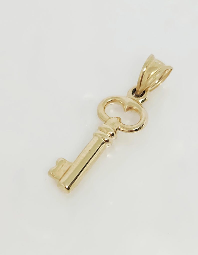 May include: A gold-colored key pendant with a classic design. The pendant features a detailed key shape with a loop at the top for a chain. The key's handle has a decorative design. The pendant is made of a shiny, reflective metal.