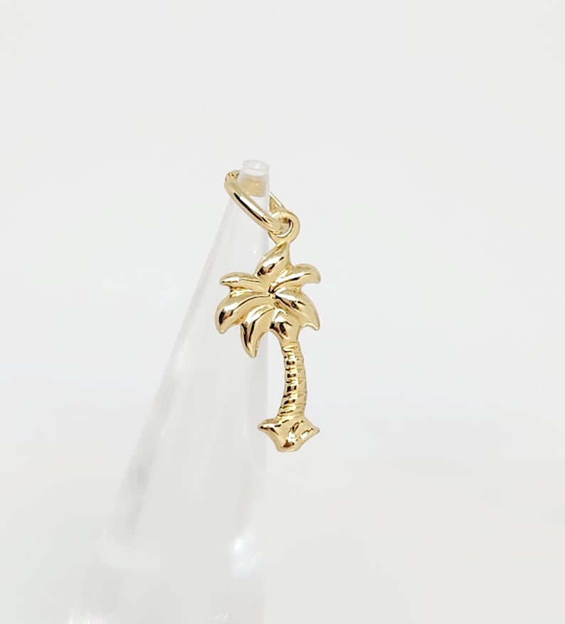 Op de afbeelding: Een gepolijste gouden bedel in de vorm van een palmboom, met een getextureerde stam en gladde, gebogen bladeren. De bedel is voorzien van een kleine ronde ring aan de bovenkant voor bevestiging en heeft een heldere, reflecterende metalen afwerking.