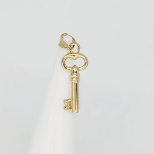 May include: A gold-colored key pendant with a classic design. The key features a detailed bow and a long, slender stem with a rectangular bit. The pendant is suspended from a small loop, ready to be added to a necklace or bracelet.