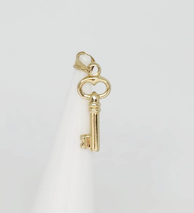 May include: A gold-colored key pendant with a classic design. The key features a detailed bow and a long, slender stem with a rectangular bit. The pendant is suspended from a small loop, ready to be added to a necklace or bracelet.