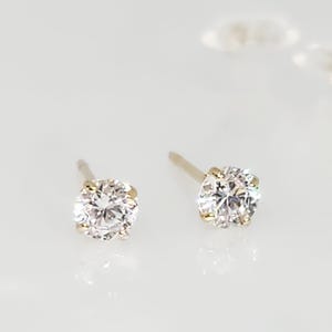 May include: A pair of stud earrings with round, clear gemstones set in gold-colored metal. The earrings are displayed on a white surface, showcasing their sparkle and design. The studs are a classic jewelry item.