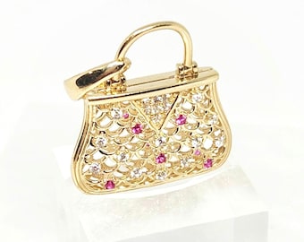 May include: A gold charm shaped like a miniature handbag featuring an intricate openwork lattice design. The surface is embellished with small, sparkling clear stones and vibrant pink stones set within the gold metal filigree pattern.