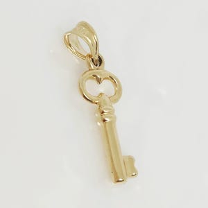 May include: A gold-colored key pendant with a heart-shaped design on the key head. The pendant is suspended from a small loop at the top. The key's shaft is detailed with vertical lines. The pendant is likely made of gold.