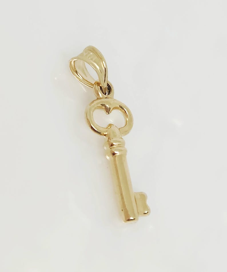 May include: A gold-colored key pendant with a heart-shaped design on the key head. The pendant is suspended from a small loop at the top. The key's shaft is detailed with vertical lines. The pendant is likely made of gold.