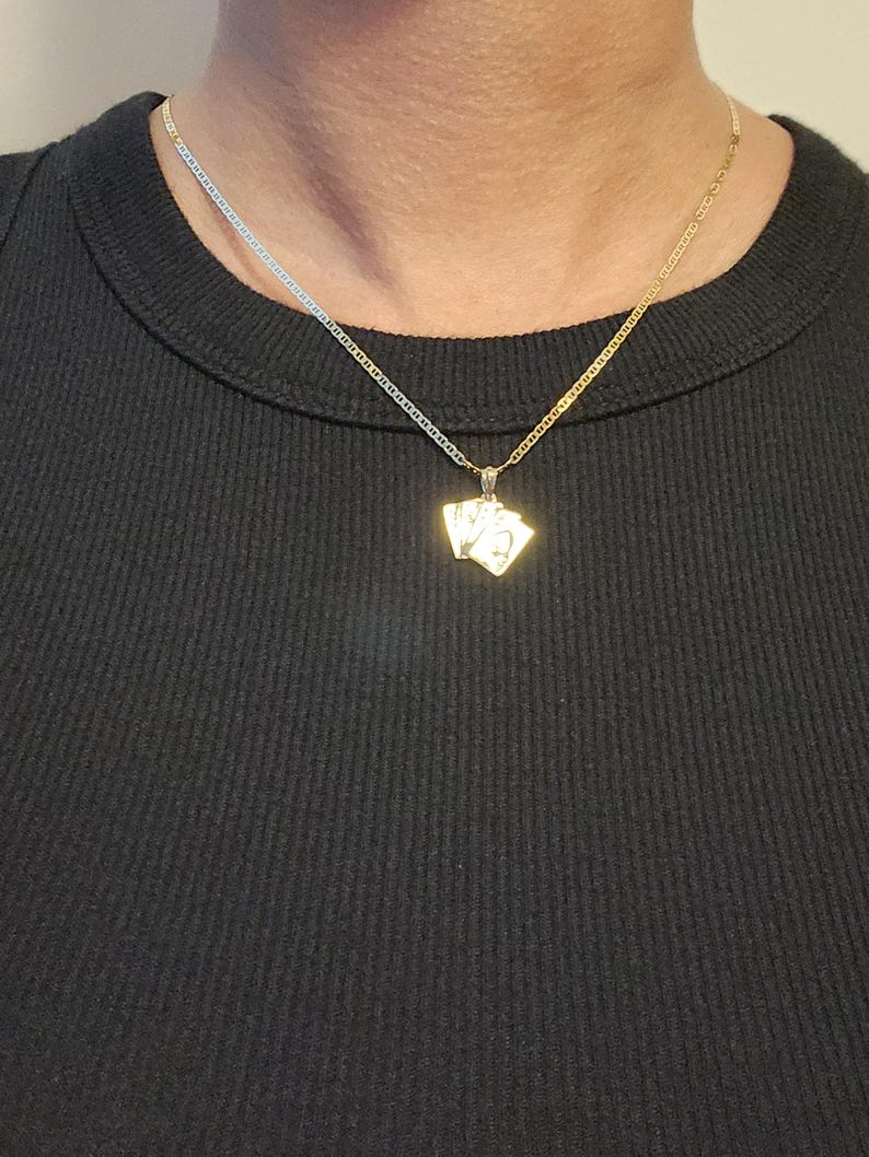 May include: A gold-toned necklace featuring a pendant shaped like a small fan of three playing cards. The pendant hangs from a delicate, thin gold-toned chain. The playing card design is metallic gold with visible engraved details on the surface.
