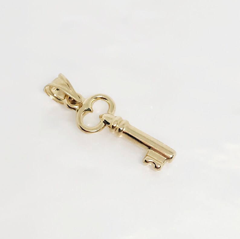 May include: A gold-colored key pendant with a classic design. The key features a detailed bow, a textured shaft, and a decorative bit. The pendant is likely made of gold and is designed to be worn as jewelry.