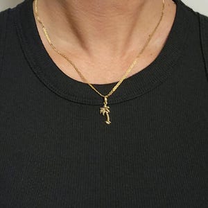 Op de afbeelding: Een goudkleurige ketting met een fijne schakelketting en een kleine, gedetailleerde palmboomhanger. De hanger hangt centraal aan de ketting, die rust op een zwart geribbelde tanktop. Het metaal heeft een gepolijste, reflecterende afwerking.