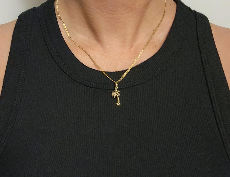 Op de afbeelding: Een goudkleurige ketting met een fijne schakelketting en een kleine, gedetailleerde palmboomhanger. De hanger hangt centraal aan de ketting, die rust op een zwart geribbelde tanktop. Het metaal heeft een gepolijste, reflecterende afwerking.