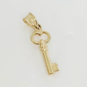 May include: A gold-colored key pendant with a heart-shaped detail in the bow. The key has a detailed barrel and bit. The pendant is designed to be worn as a necklace charm.