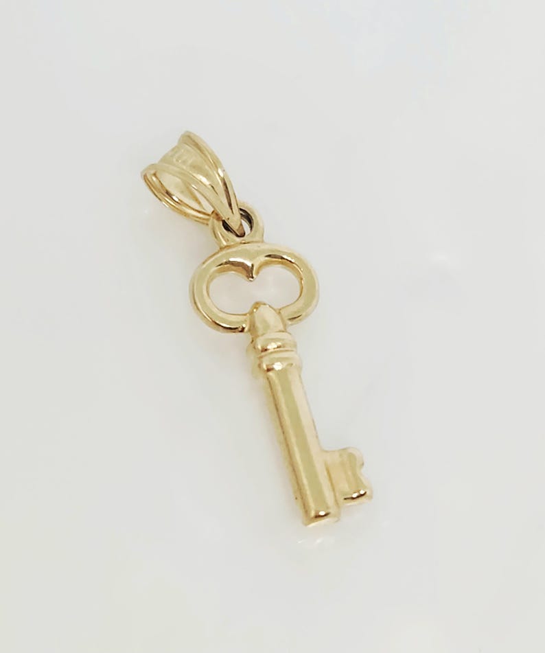 May include: A gold-colored key pendant with a heart-shaped detail in the bow. The key has a detailed barrel and bit. The pendant is designed to be worn as a necklace charm.