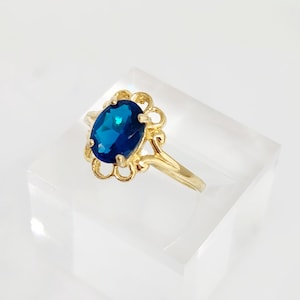 May include: A gold-colored ring featuring a large, oval-cut, deep blue gemstone. The gemstone is set in a decorative, openwork setting. The ring is displayed on a clear, angled surface against a white background.