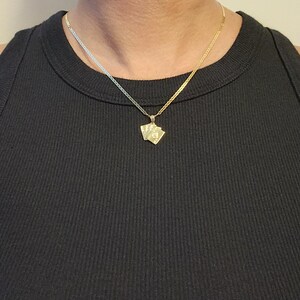 May include: A gold-toned necklace featuring a pendant shaped like a small fan of playing cards. The pendant hangs from a delicate, flat-link chain that rests against the neck. The metal has a polished, reflective finish that catches the light.