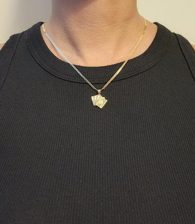 May include: A gold-toned necklace featuring a pendant shaped like a small fan of playing cards. The pendant hangs from a delicate, flat-link chain that rests against the neck. The metal has a polished, reflective finish that catches the light.