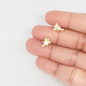 Puede incluir: Dos peque&ntilde;os pendientes de bot&oacute;n dorados con forma de flor. Los pendientes se sostienen en la mano de una persona sobre un fondo blanco. Los pendientes tienen un dise&ntilde;o floral detallado.