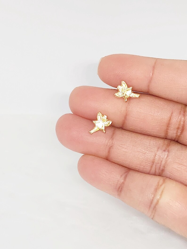Puede incluir: Dos peque&ntilde;os pendientes de bot&oacute;n dorados con forma de flor. Los pendientes se sostienen en la mano de una persona sobre un fondo blanco. Los pendientes tienen un dise&ntilde;o floral detallado.