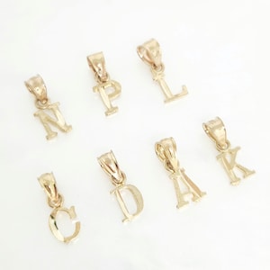 Puede incluir: Una colección de siete colgantes individuales con forma de letra en metal dorado, cada uno con un acabado pulido y una pequeña anilla integrada en la parte superior para su fijación. Las letras mostradas son N, P, L, C, D, A y K, dispuestas en dos filas.