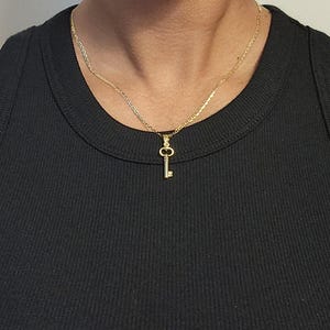 May include: A gold-colored key pendant necklace. The key charm hangs from a delicate chain. The necklace is worn against a black ribbed tank top.