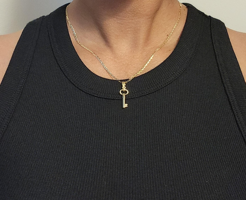 May include: A gold-colored key pendant necklace. The key charm hangs from a delicate chain. The necklace is worn against a black ribbed tank top.