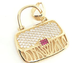 May include: A gold-toned pendant shaped like a miniature handbag, featuring a top flap encrusted with numerous clear white stones and a single rectangular red stone at the center. The lower portion of the bag has a textured, open-work design.