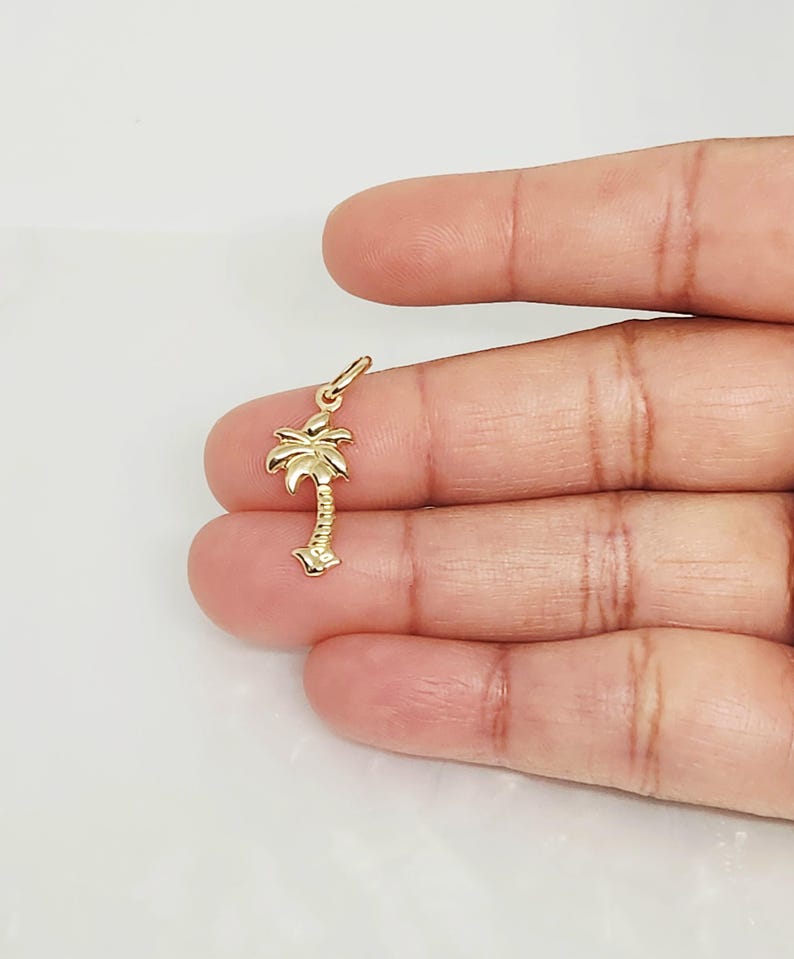 Op de afbeelding: Een kleine hanger van gepolijst geelgoud in de vorm van een palmboom. De hanger heeft een gestructureerde stam en gedetailleerde bladeren aan de bovenkant, met een kleine ronde ring aan de bovenzijde om aan een ketting of armband te bevestigen.