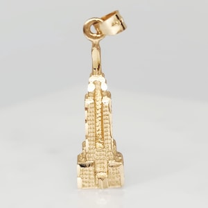 May include: A gold-colored Empire State Building charm. The charm is a miniature replica of the iconic skyscraper, featuring detailed architectural elements. The charm is designed to be attached to a necklace or bracelet.