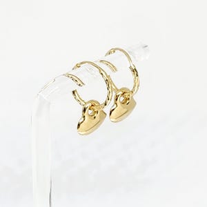 May include: A pair of gold hoop earrings featuring a textured, twisted metal design on the hoops and a smooth, polished heart-shaped charm dangling from each hoop. The earrings are displayed on a clear, curved stand against a white background.