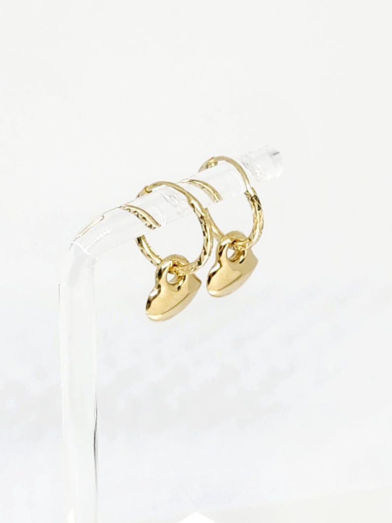 May include: A pair of gold hoop earrings featuring a textured, twisted metal design on the hoops and a smooth, polished heart-shaped charm dangling from each hoop. The earrings are displayed on a clear, curved stand against a white background.