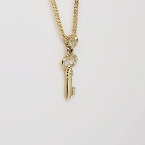 May include: A gold-colored key pendant hangs from a delicate gold chain. The key pendant is detailed with a classic key design. The chain and pendant are displayed against a white background.