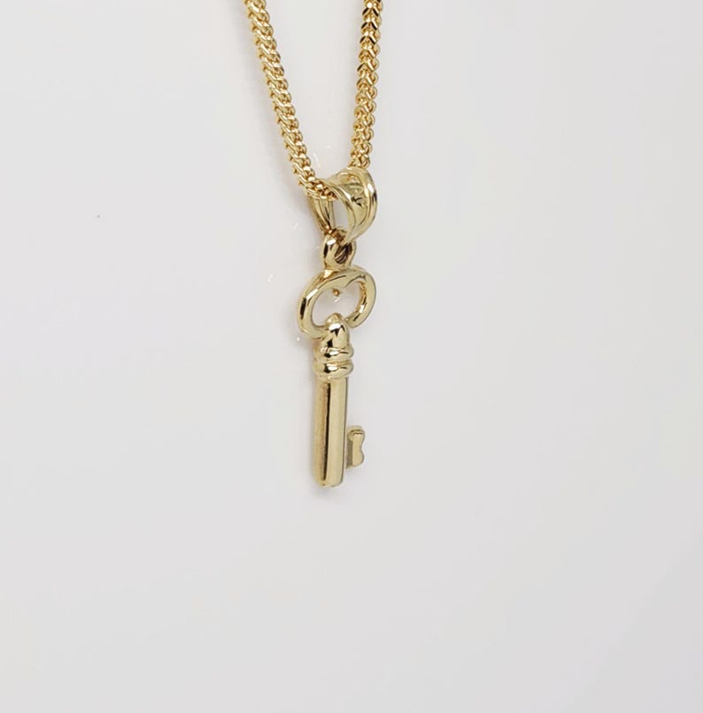 May include: A gold-colored key pendant hangs from a delicate gold chain. The key pendant is detailed with a classic key design. The chain and pendant are displayed against a white background.