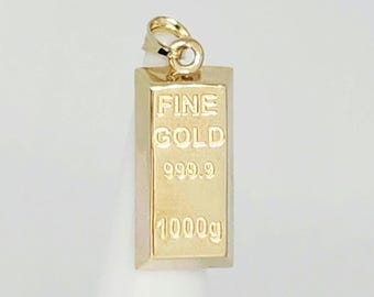 May include: A gold-colored metal pendant shaped like a rectangular bullion bar. The front surface features embossed text reading FINE GOLD, 999.9, and 1000g. A small circular jump ring is attached to the top of the bar for use as a jewelry charm.