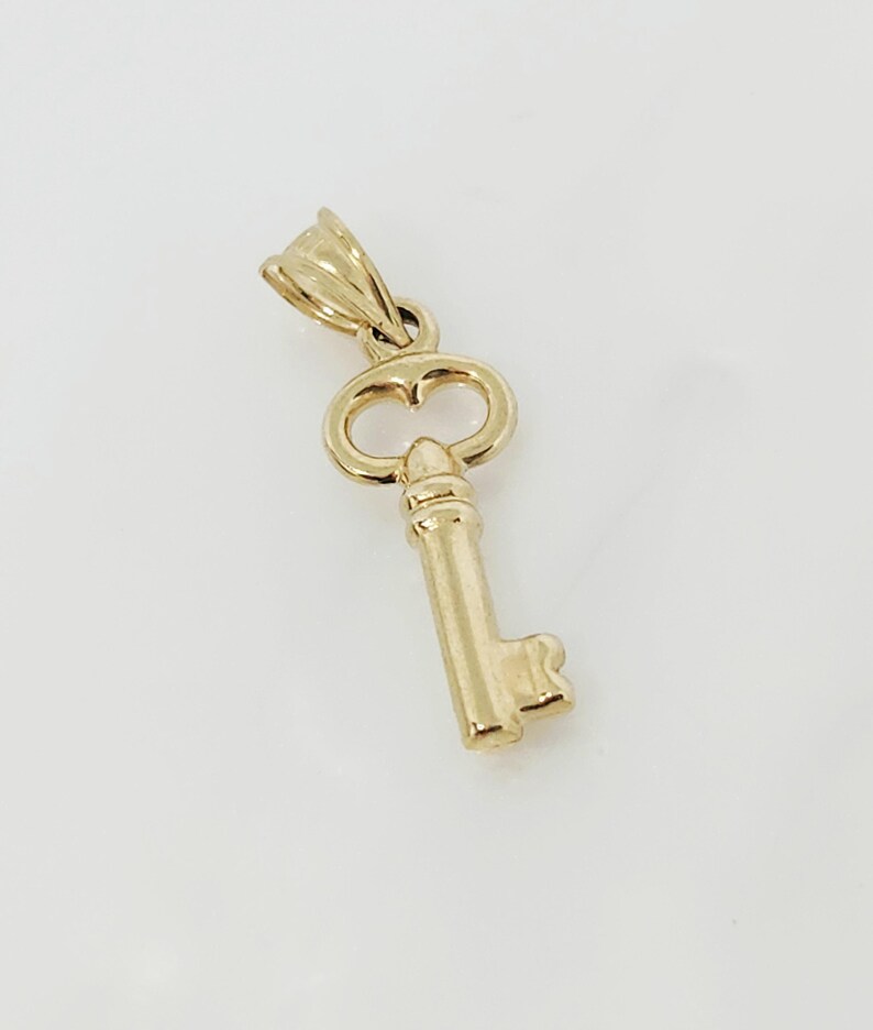 May include: A gold-colored key pendant with a detailed design. The key has a classic shape with a loop at the top for a chain. The pendant is made of a shiny, reflective metal.