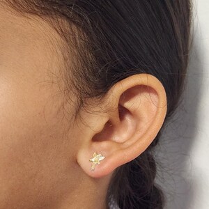 Puede incluir: Un pendiente de bot&oacute;n dorado con forma de hada se lleva en una oreja. El pendiente tiene un dise&ntilde;o detallado, con alas visibles y una forma delicada. La oreja se encuentra sobre un fondo neutro, resaltando la joya.