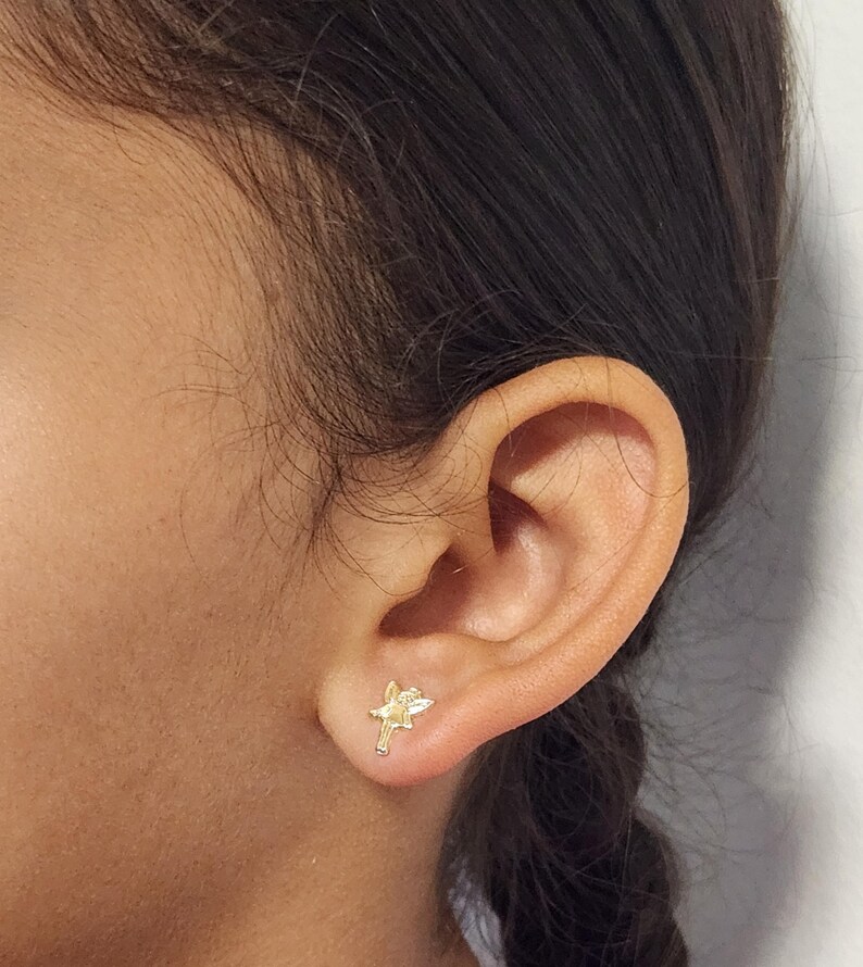 Puede incluir: Un pendiente de bot&oacute;n dorado con forma de hada se lleva en una oreja. El pendiente tiene un dise&ntilde;o detallado, con alas visibles y una forma delicada. La oreja se encuentra sobre un fondo neutro, resaltando la joya.